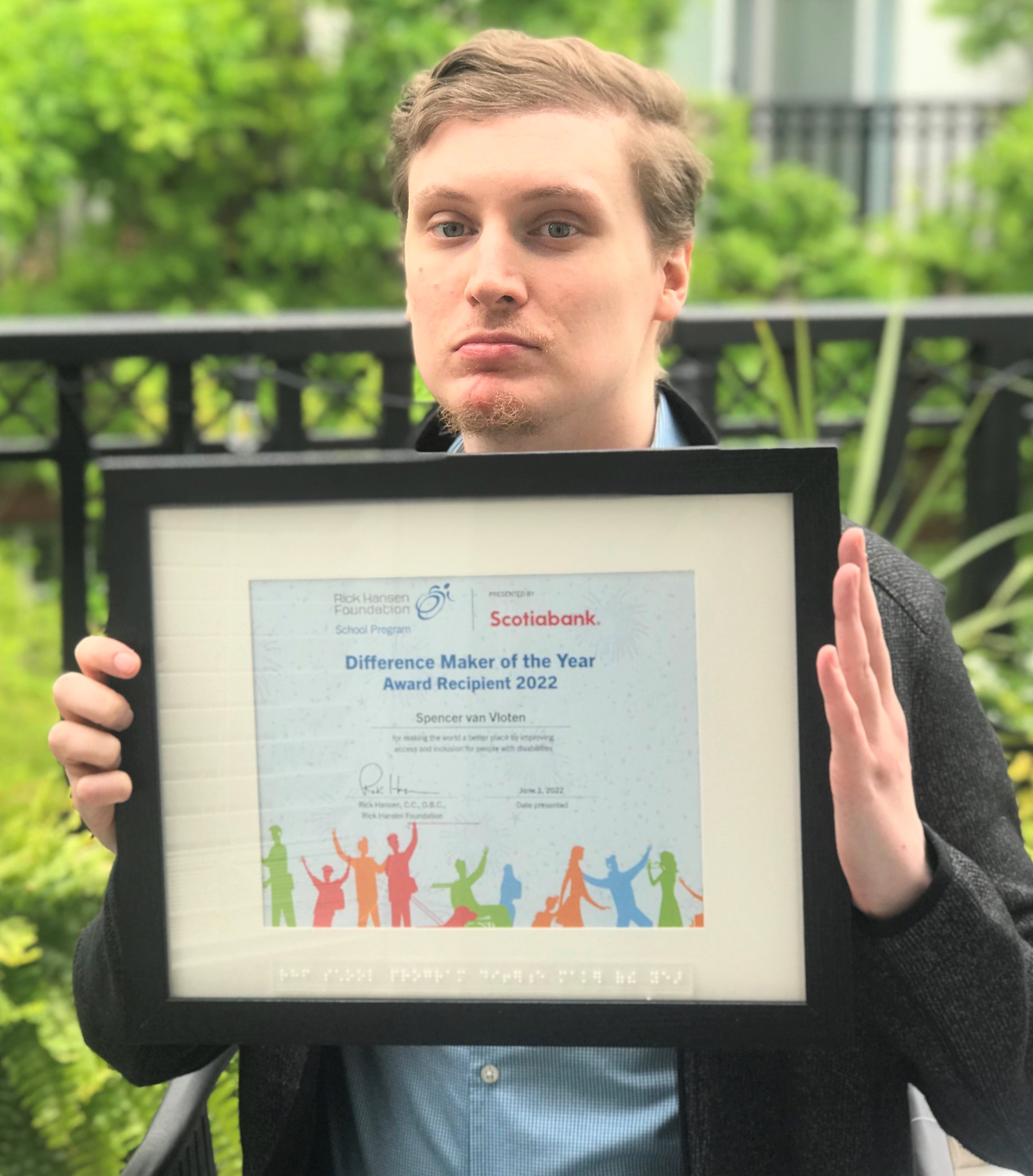 Spencer, who has short brown hair and light-coloured eyes, holding his Difference Maker certificate in front of a tree-filled background.